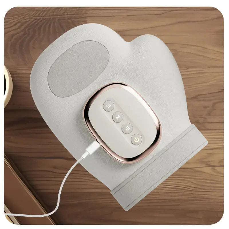 Luliqua HandRelief Pro white hand massager with rose gold trim charging on white hand-shaped charging mat - top view of white rounded rectangular massager with rose gold metallic border, 4 control buttons showing dollar sign, mode, intensity and power icons, white USB charging cable connected, placed on white silicone hand-shaped charging mat with oval cutout for massager, natural wood grain table surface